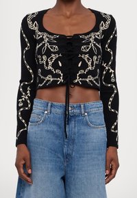 Haut noir à manches longues court avec motifs floraux crème et détail à lacets sur le devant, porté avec un jean bleu clair taille haute.