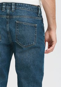 Dunkelblaue Jeans aus Denim mit geradem Schnitt, ausgestattet mit zwei Gesäßtaschen und einem sichtbaren Bund mit Gürtelschlaufen. Strukturiertes Stoffdetail.