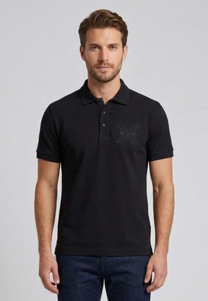 Uomo in piedi che indossa una polo nera con un discreto logo "Richmond X" sul petto e jeans blu scuro.