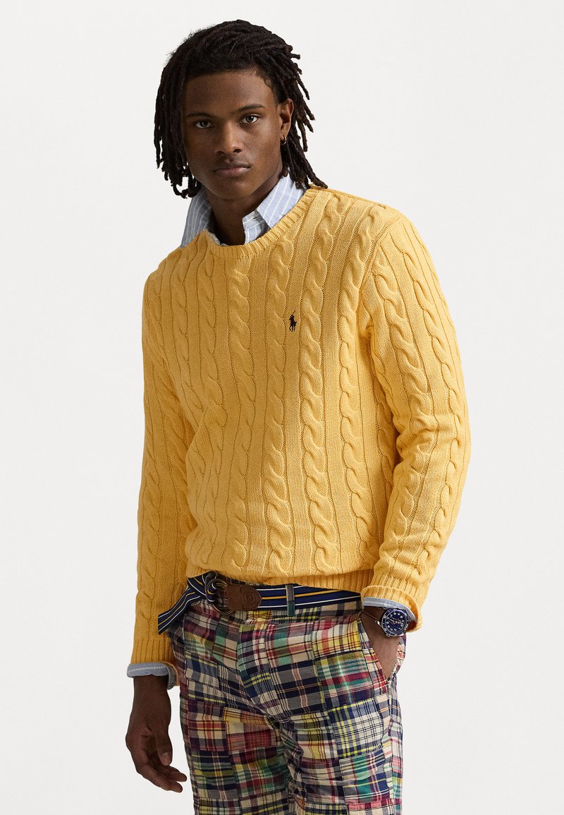 Un pull en tricot jaune à col en V, superposé sur une chemise à col bleu clair. Assorti à un pantalon à carreaux multicolores et une ceinture marron.