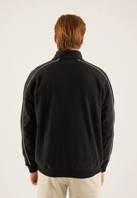 Mand med lysbrunt hår iført en sort fleecejakke med hvid piping langs ærmerne og beige bukser, der står med ryggen til mod en ensfarvet baggrund.