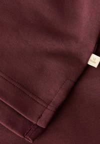 Bordeauxroter Baumwollstoff mit glatter Textur, versehen mit abgesteppten Nähten und einem kleinen Markenetikett in einem helleren Cremeton.