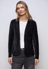 Jeune femme portant un blazer en velours côtelé noir sur une chemise blanche et un jean foncé, debout devant un fond clair uni.
