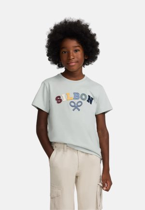 Camiseta de algodón azul claro con "SILBON" bordado en letras multicolores. Mangas cortas, escote redondo, combinada con pantalones cargo beige.