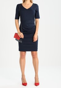 Marinblå knä-lång klänning med V-ringning och korta ärmar, med inslag av veckade sidodetaljer. Parat med en röd och svart handväska och röda klackar.