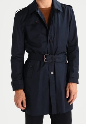 Homme portant un trench-coat bleu marine ceinturé sur un pull marron, debout devant un fond clair uni.