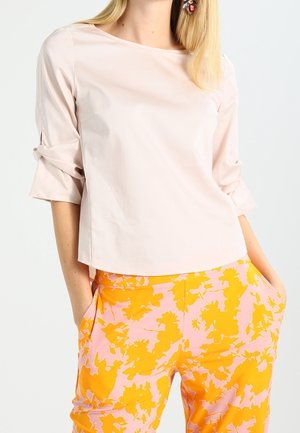 Mujer con blusa de manga tres cuartos de color rosa pálido y pantalones de talle alto rosa con un brillante patrón floral amarillo, con las manos en los bolsillos.