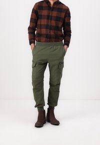 Brun och svart rutigt knapp-down skjorta, gröna cargo byxor med elastisk midja, dragkedjor och fickor, tillsammans med bruna ankelstövlar.