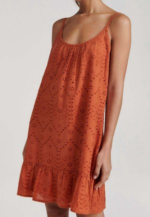 Femme portant une robe sans manches orange avec des motifs floraux ajourés et un ourlet à volants, posant devant un fond neutre.