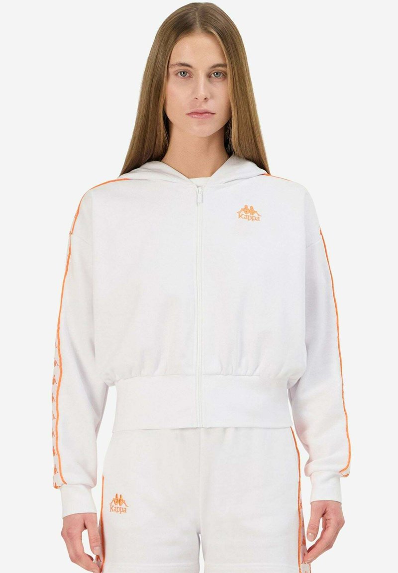 Sudadera blanca con cremallera y detalles en naranja, incluyendo un logo. Presenta capucha, cinturilla elástica y franjas laterales con una marca repetida. Material suave.