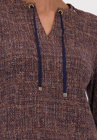 Bruine textuurstof top met een V-hals, verfraaid met gouden ogen en marineblauwe koorden, met een geweven patroon door het hele kledingstuk.