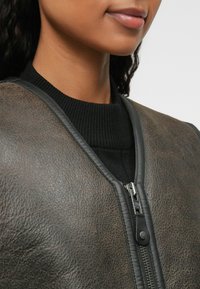 Bruine leren bomberjack met een ribbelachtige zwarte kraag en een metalen rits met leren trekband. Getextureerd oppervlak met subtiele variaties.