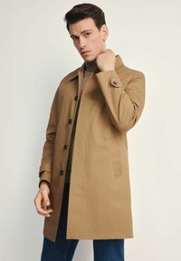 Khaki trencz z kołnierzem, zapinany na guziki, z bocznymi kieszeniami. Noszony na brązowym swetrze, prezentowany na jasnym tle.