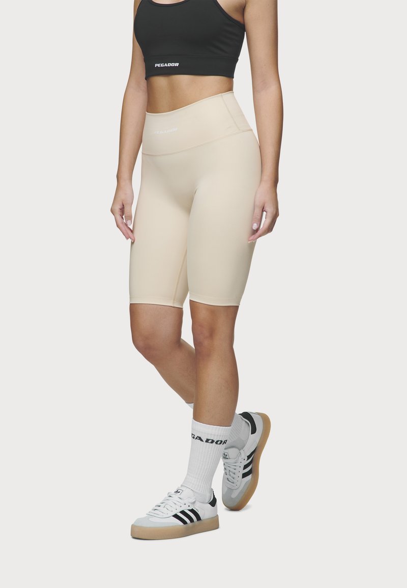 Beige, magas derekú kerékpáros rövidnadrág, rugalmas anyagból, szoros illeszkedéssel és sima textúrával, fekete crop top-pal és sportcipővel párosítva.