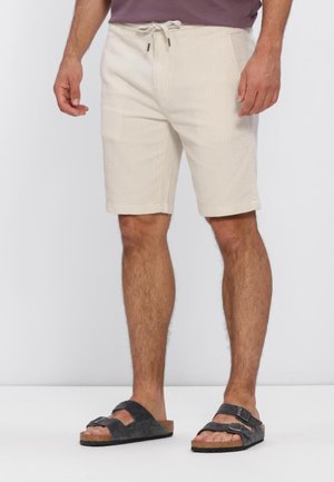 Mies, jolla on beige nauhalla kiristettävät shortsit ja mustat kaksinkertaiset remmisandaalit, seisoo yksinkertaisen vaalean taustan edessä.