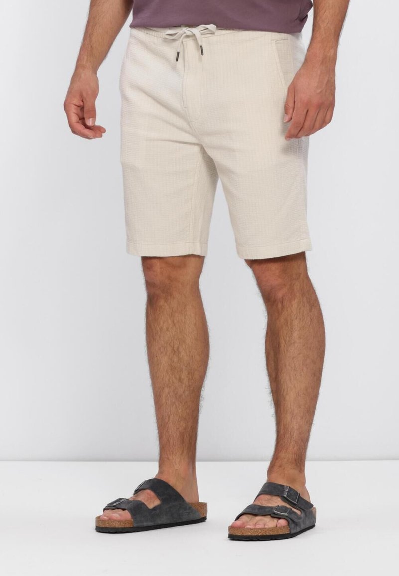 Homme portant un short beige avec cordon et des sandales noires à double bride, debout devant un fond clair uni.
