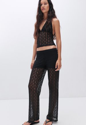 Set in pizzo nero composto da due pezzi, con top corto a scollo a V e pantaloni a gamba larga con design trasparente. Tessuto morbido e testurizzato con motivi floreali.