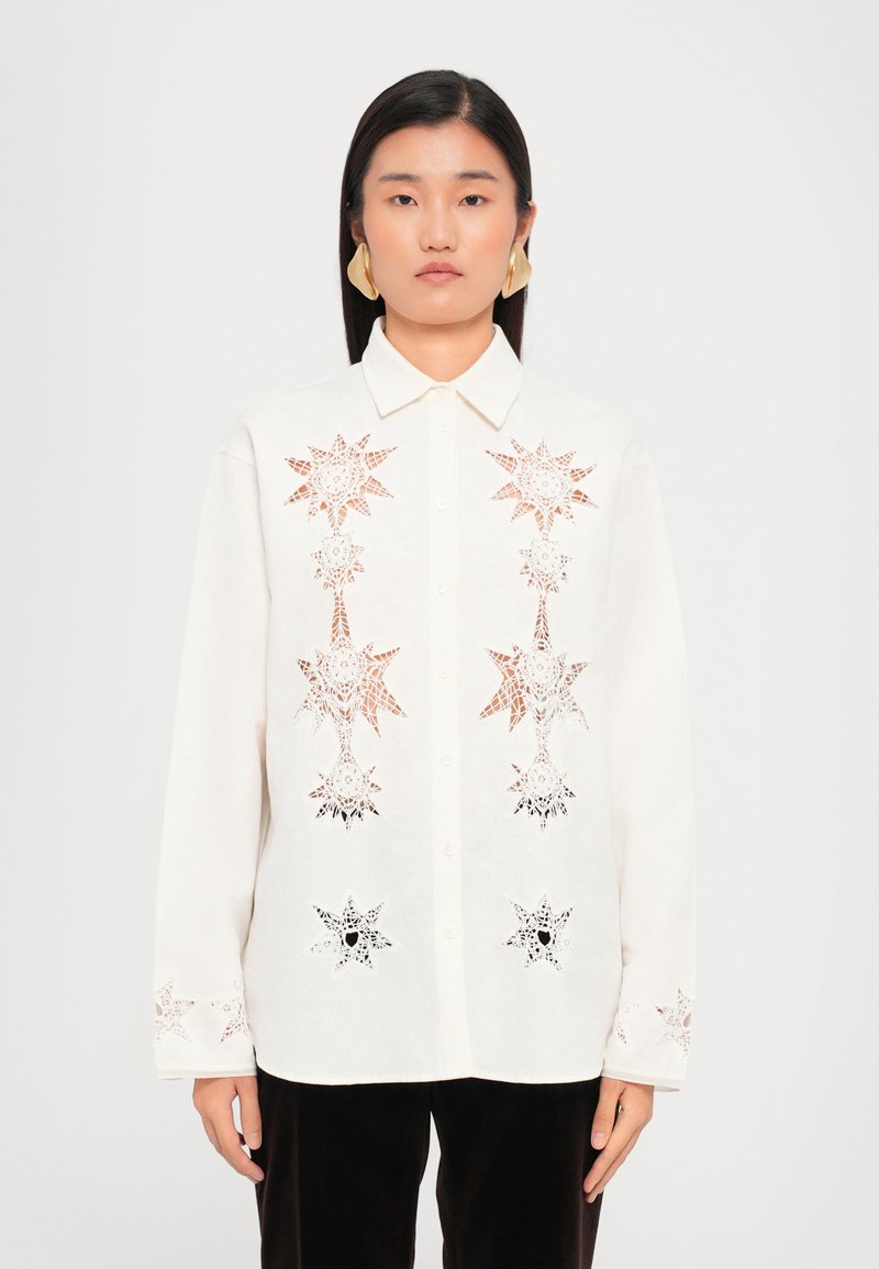 White shirt with button-up front, features star-shaped cutouts in red and black lace, long sleeves with similar detailing, and a collar.