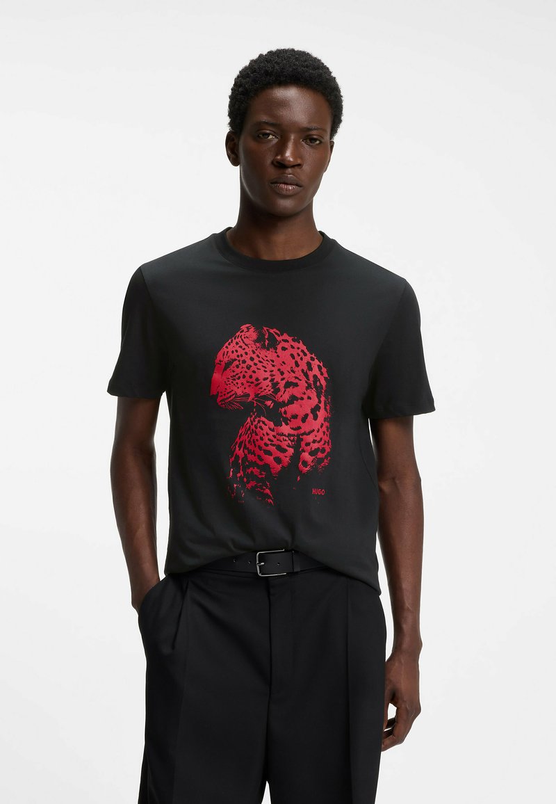 Homme portant un t-shirt noir avec un motif léopard rouge, ceinture noire et pantalon noir, debout devant un fond blanc uni.