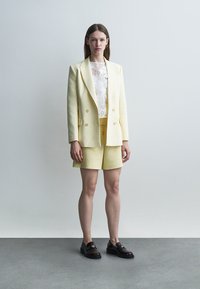 Žena oblečená v bledozltom saku a kraťasoch s bielym čipkovaným topom a čiernymi mokasínami, stojaca na obyčajnom sivom pozadí.