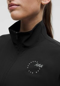 Crna jakna na zip s visokim ovratnikom, od teksturiranog materijala, koja na lijevoj strani prsnog područja ima okrugli bijeli logo sa stiliziranim figurama.
