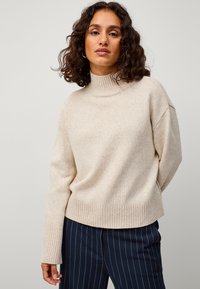 Beige coltrui met een ribgebreide zoom en boorden. Regular fit en zachte textuur. Draagt met marineblauwe gestreepte broek, gedeeltelijk zichtbaar.