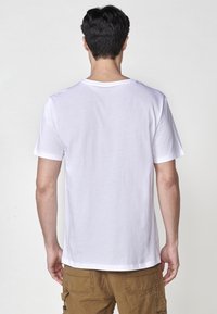 T-shirt bianco a maniche corte con collo rotondo, realizzato in cotone. Vista da dietro, abbinato a pantaloni cargo beige. Design semplice, senza motivi.