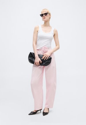 Γυναίκα με λευκό τοπ χωρίς μανίκια και φαρδιά ροζ παντελόνια με floral ζώνη, μαύρες μυτερές γόβες, μεγάλο φάκελο τσάντας και μαύρα γυαλιά ηλίου.