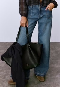 Bolso tote de cuero negro con un acabado texturizado, de gran tamaño y asas verdes. Un suéter de punto azul marino está apoyado sobre el bolso.