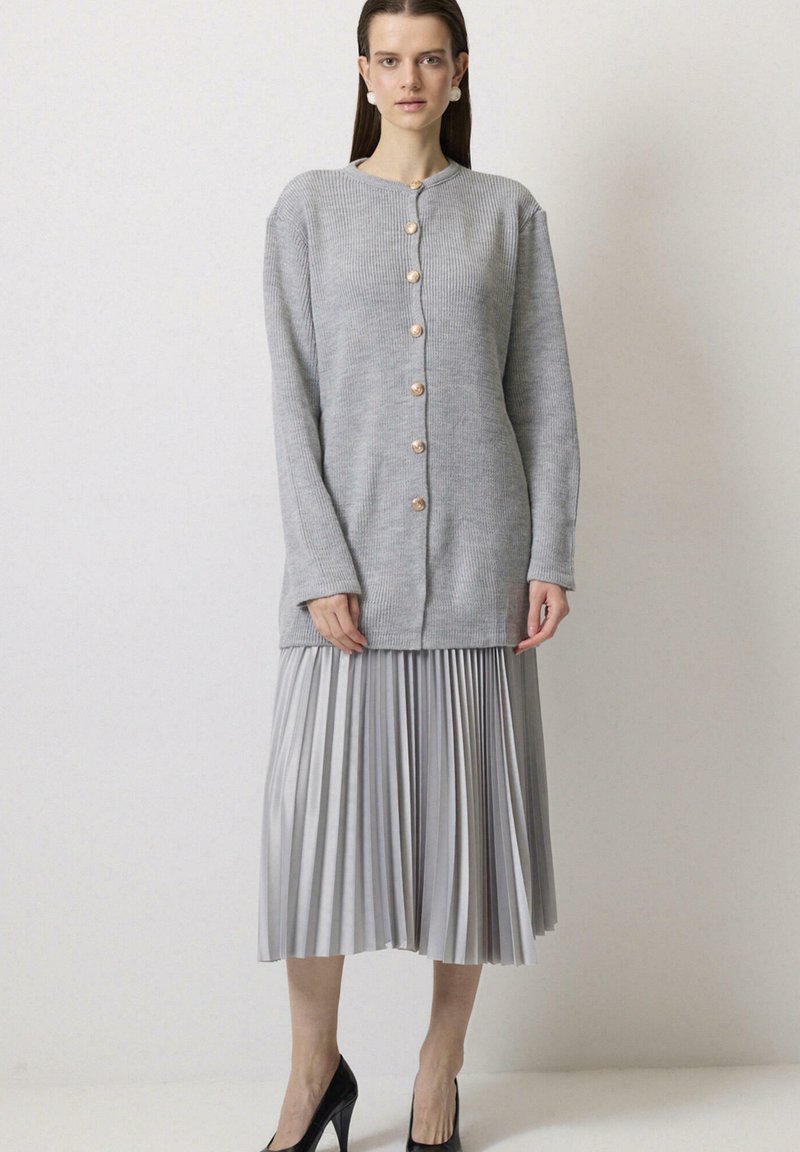 Szary prążkowany kardigan z dużymi różowymi złotymi guzikami, połączony z plisowaną srebrną spódnicą. Czarne szpilki dopełniają strój.
