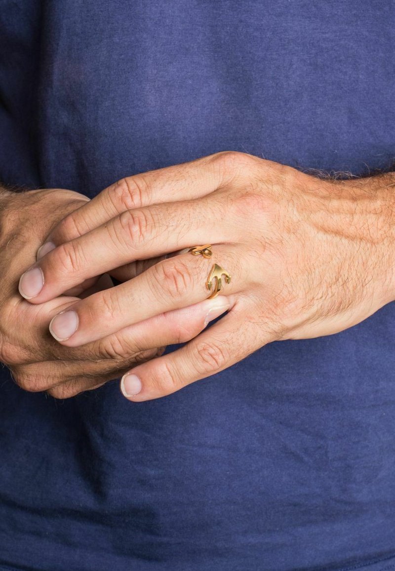 Anello d'oro con un design ondulato, indossato su un dito. Le mani sono leggermente abbronzate con texture della pelle visibile, su uno sfondo di camicia blu.
