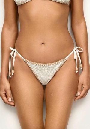 Kobieta w białych teksturowanych spodniach od bikini z bocznymi wiązaniami i drewnianymi koralikami, pokazana od talii do połowy uda na jednolitym tle.