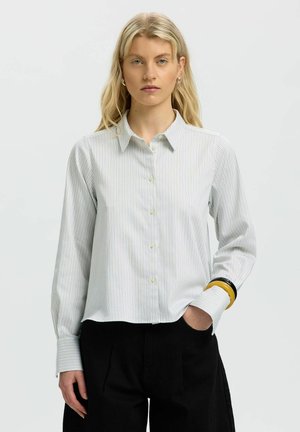 Jeune femme portant une chemise claire à rayures boutonnée et un pantalon noir, debout avec une main dans la poche sur un fond uni.