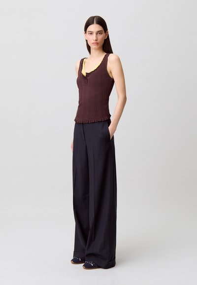 Canottiera marrone a coste con rifinitura gialla, caratterizzata da un dettaglio con bottoni. Abbinata a pantaloni larghi blu navy a righe sottili. Texture liscia, vestibilità rilassata.
