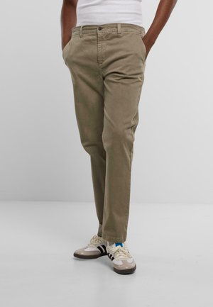 Pantalones chinos beige de corte clásico, con bolsillos frontales, cremallera y tela suave. Combinados con zapatillas deportivas y una camiseta sin mangas blanca ajustada.