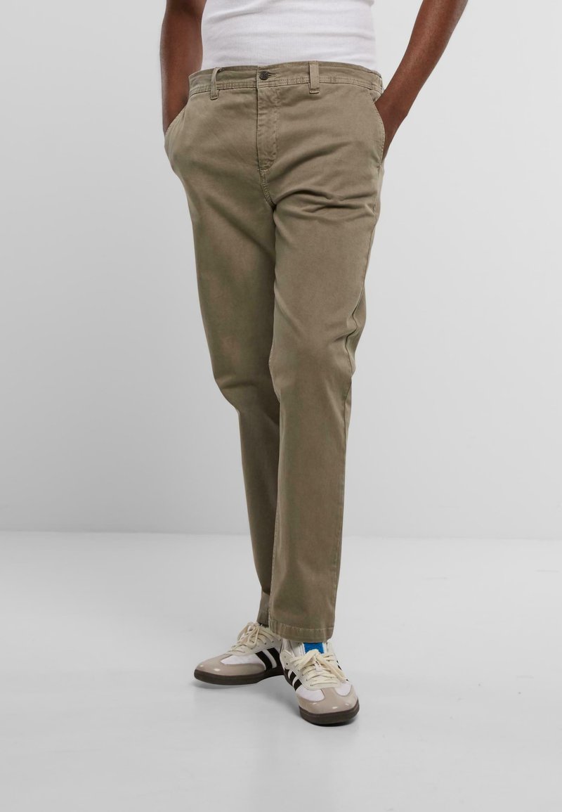 Pantalones chinos beige de corte clásico, con bolsillos frontales, cremallera y tela suave. Combinados con zapatillas deportivas y una camiseta sin mangas blanca ajustada.