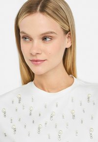 Top bianco con scollatura rotonda, decorato con perlini finti e gemme scintillanti sparse, caratterizzato da una texture di tessuto liscia.