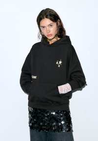 Zwarte hoodie met gouden steraccenten en geborduurde tekst, met kanten manchetten over een zwart paillettenkleed. Casual, losse pasvorm.