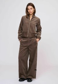 Barna kordbársony dzseki magas gallérral és elülső cipzárral, széles szárú nadrággal azonos anyagból. Mindkét darab oldalzsebekkel rendelkezik.