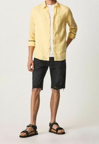 Camisa de lino amarillo claro con mangas enrolladas, camiseta blanca debajo, pantalones cortos de mezclilla negra desgastados y sandalias negras con suelas de corcho.