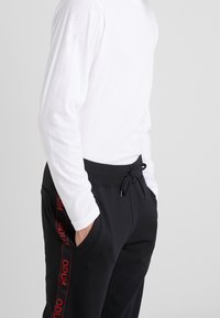 Schwarze Jogginghosen mit einem Kordelzugbund und rotem Branding entlang der Seite. Kombiniert mit einem weißen Langarmshirt. Glatte Textur.