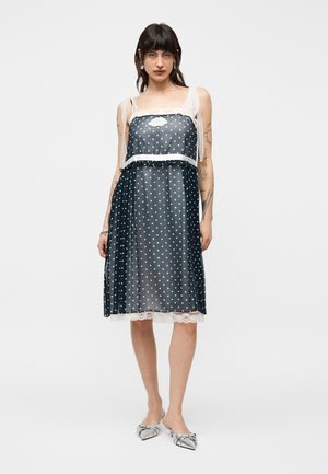 Donna con indosso un abito nero senza maniche a pois bianchi, bordato di pizzo, con spalline bianche trasparenti, che posa con scarpe piatte argento con punta appuntita.