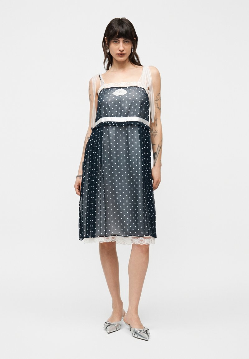 Femme portant une robe noire sans manches à pois blancs, ornée de dentelle et de bretelles blanches transparentes, posant avec des ballerines argentées à bout pointu.