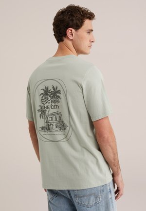 Mann, seitlich stehend, trägt ein hellgrünes T-Shirt mit schwarzen Palmen, einem Gebäude und dem Schriftzug „Escape The City“ auf dem Rücken.