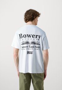 Balta medvilnės marškinėliai su dideliu juodu tekstu nugaroje: "Bowery ATELIER Lower East Side" ir "ORIGINALS™ STUDIO." Apskritas kaklas, trumpomis rankovėmis.