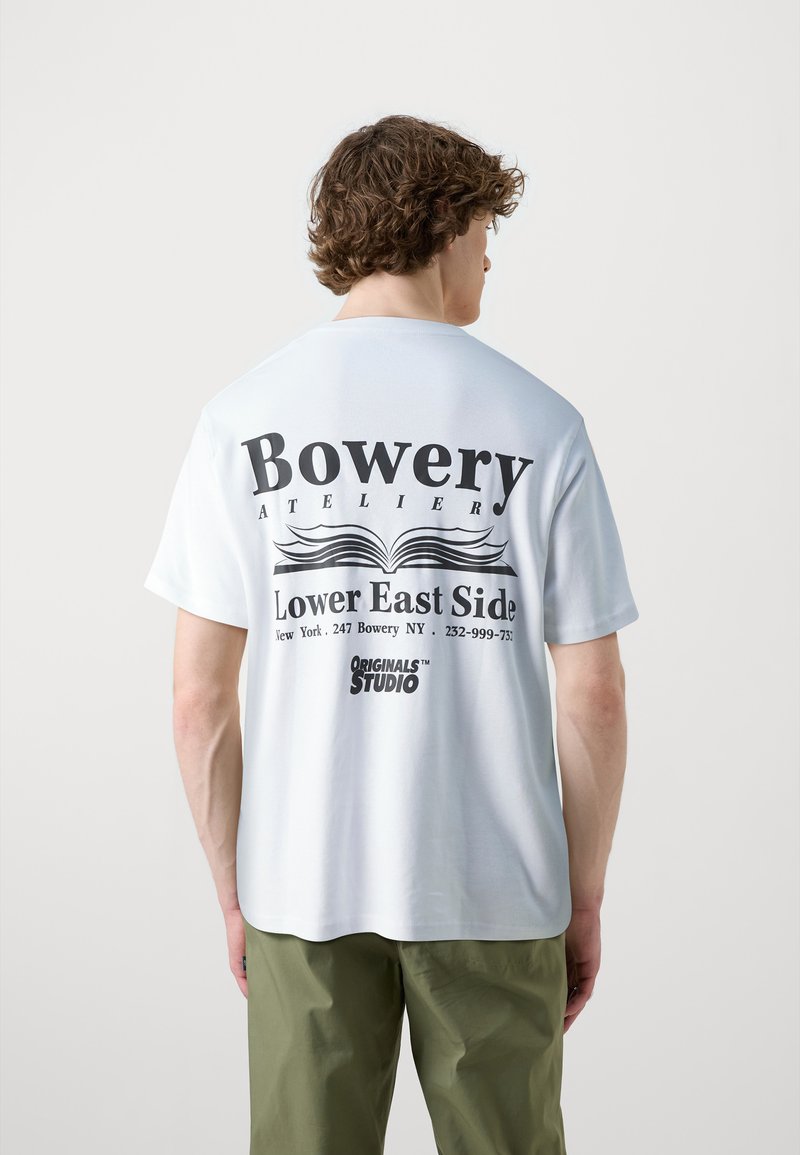 Balta medvilnės marškinėliai su dideliu juodu tekstu nugaroje: "Bowery ATELIER Lower East Side" ir "ORIGINALS™ STUDIO." Apskritas kaklas, trumpomis rankovėmis.
