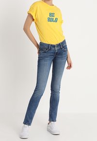 T-shirt di cotone gialla con stampa "BE BOLD", abbinata a jeans skinny blu. Il modello indossa sneaker bianche. Outfit semplice e informale.