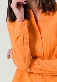 Chemise boutonnée en orange vif, fabriquée en tissu texturé. Présente un col, des manches bouffantes et des boutons brillants sur le devant et les poignets.