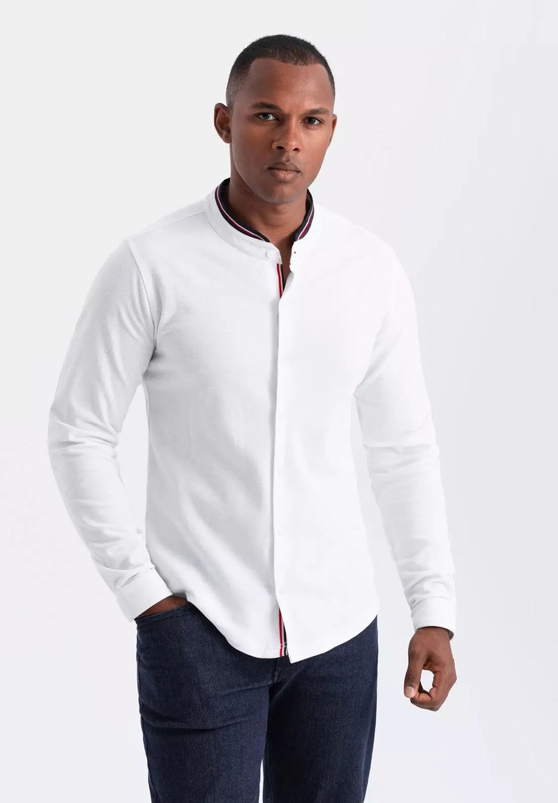 Witte blouse met lange mouwen, een staande kraag en een voorste sluiting. Heeft contrasterende strepen op de binnenkant van de kraag. Gepaard met donkerblauwe jeans.