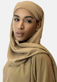 Ein Modell trägt einen beigen Hijab und passende Kleidung. Der Stoff ist glatt mit subtiler sheen, was ein einfaches Design und klare Linien zur Geltung bringt.